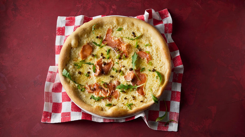 Pizza topped with smoked salmon, cheese, rocket and capers. The pizza sits on a red and white checkered cloth on a red table.