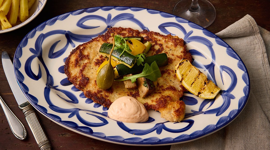 Panerad schnitzel på tallrik med blå kant, toppad med sallad, oliver och grillad citron, serverad med rosa sås.