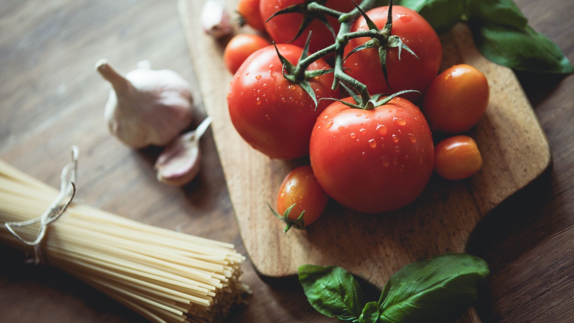 Wooden cutting board with vine tomatoes, pasta, garlic and basil.