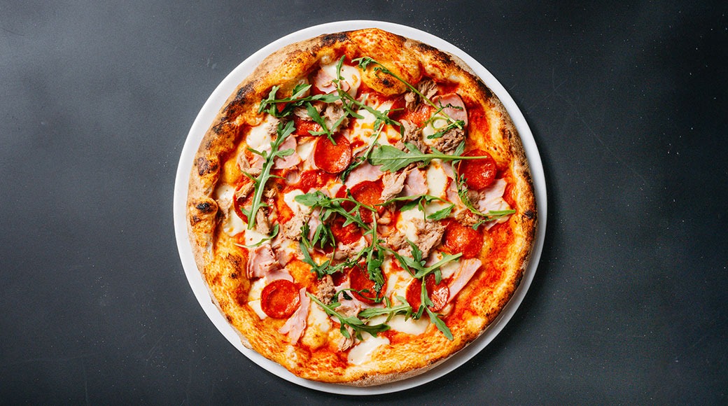 Pizza valkoisella lautasella tummalla alustalla, täytetty kinkkua, pepperonia, juustoa ja rucolaa.