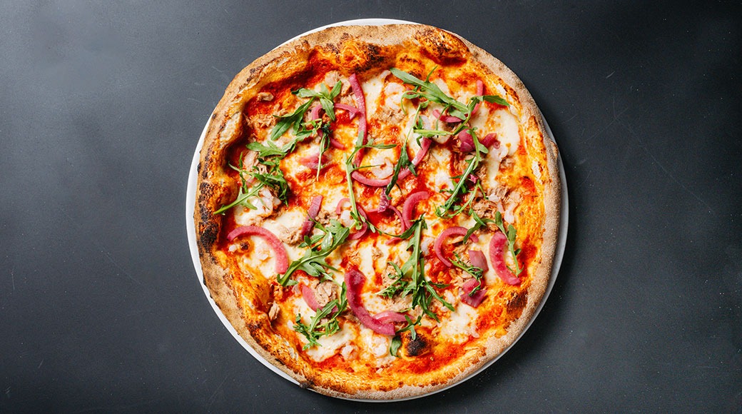 Pizza juustolla, tonnikalalla ja rucolalla tummalla alustalla.