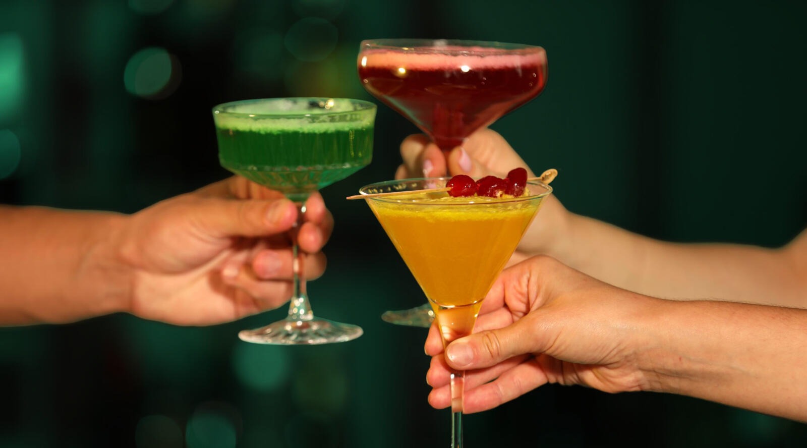 Kolme kättä pitelee kuppimaljaisia mocktaillaseja, joissa on vihreä, punainen ja keltainen juoma.