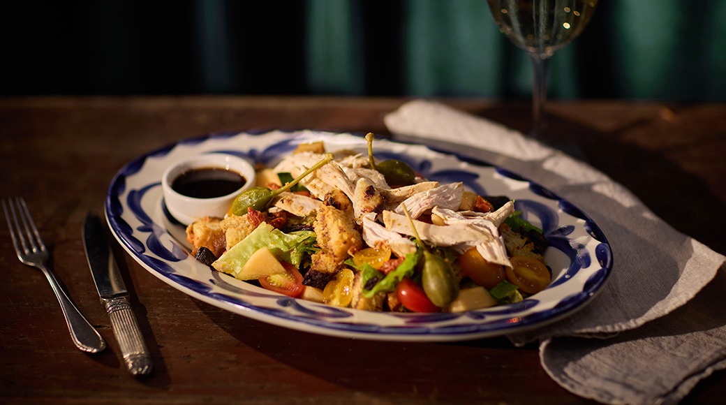 Värviline salat sini-valge äärega taldrikul kanaga, oliivide, tomatite ja kastmega. Taustal veiniklaas.