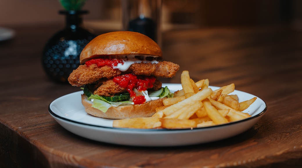 Hampurilainen täytettynä rapeilla kanafileillä, kastikkeella ja vihanneksilla. Vieressä annos ranskanperunoita valkoisella lautasella puupöydällä.