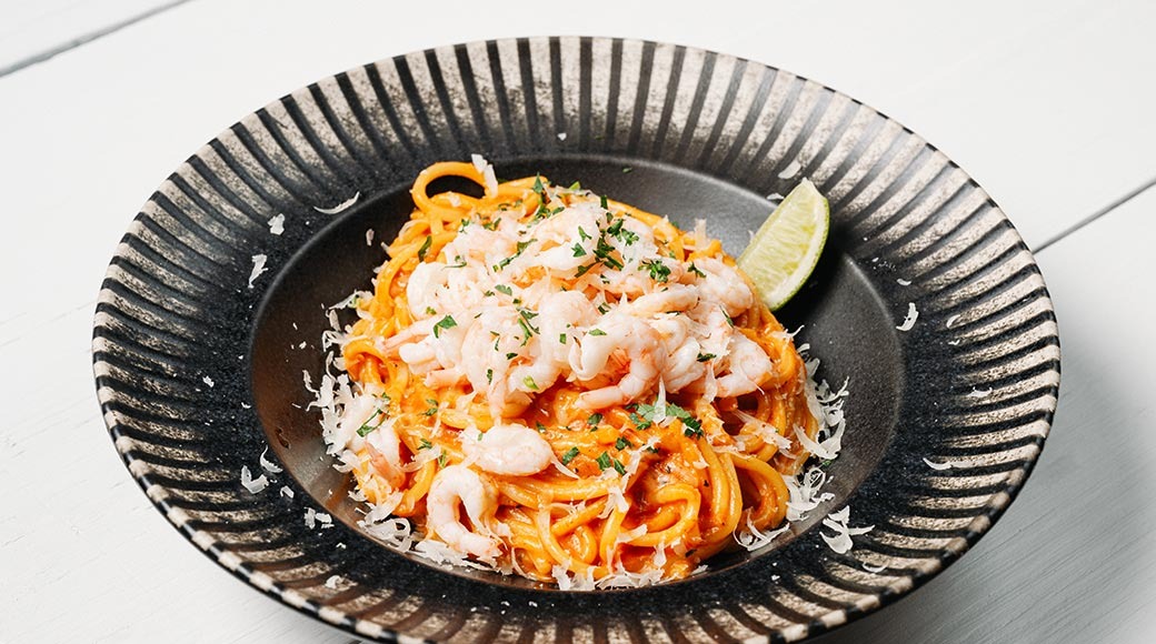 Tume triibuline taldrik krevetipastaga tomatikastmes. Peal riivitud parmesan ja ürdid, serval laimikiil.