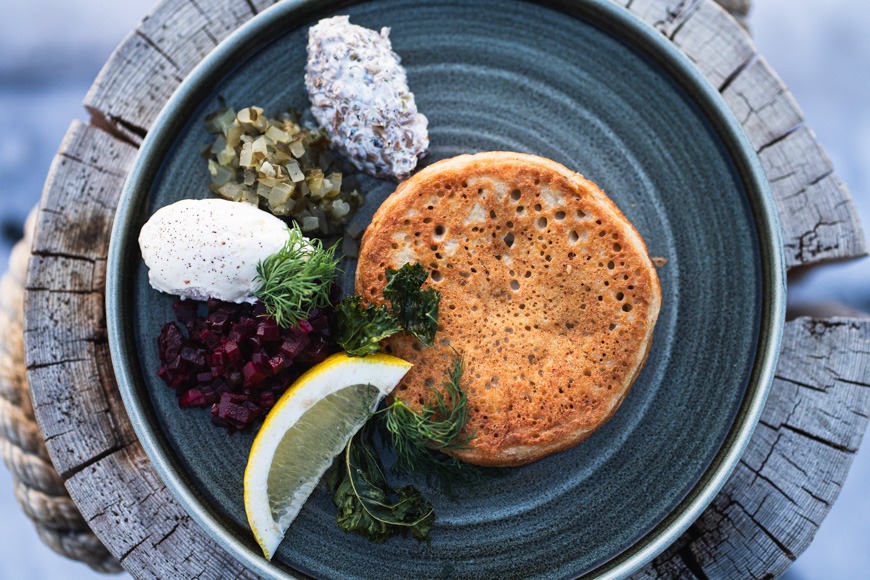 Sinisellä lautasella blini tarjoiltuna perinteisillä lisukkeilla: smetanaa, mätiä, punajuurisalaattia, yrttejä ja sitruunalohko puisella alustalla.