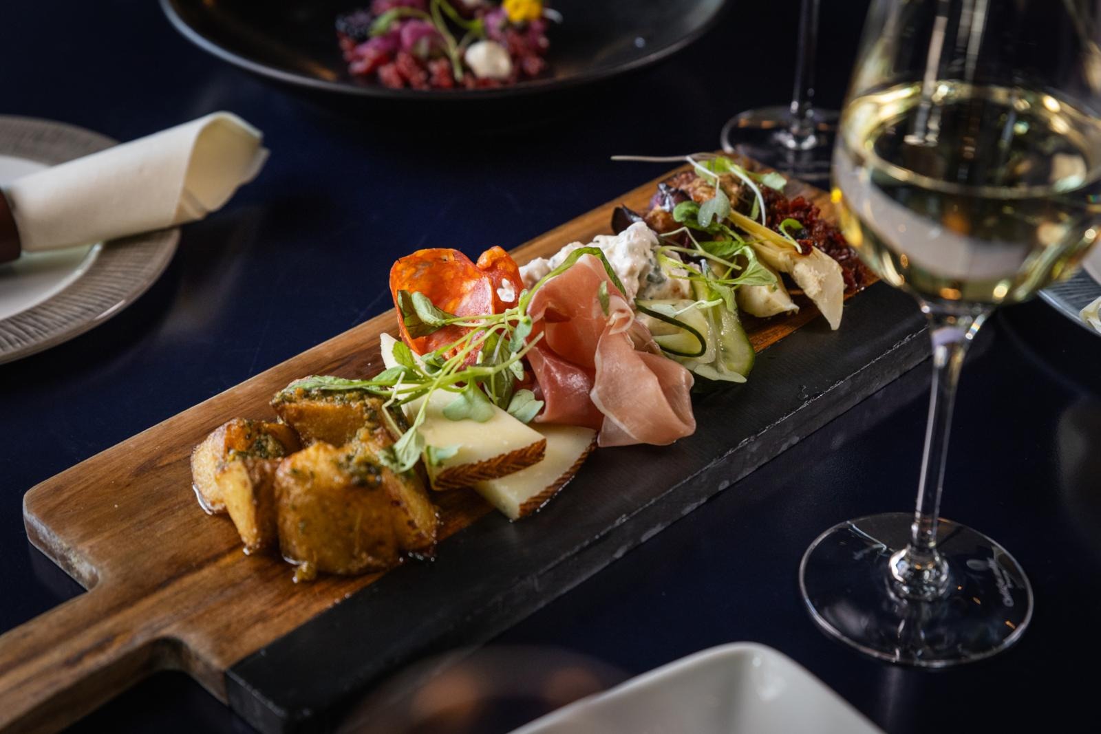 En antipastabricka serverad på skärbräda med ostar, charkuterier och rostade brödskivor. Bredvid står ett glas vitt vin.