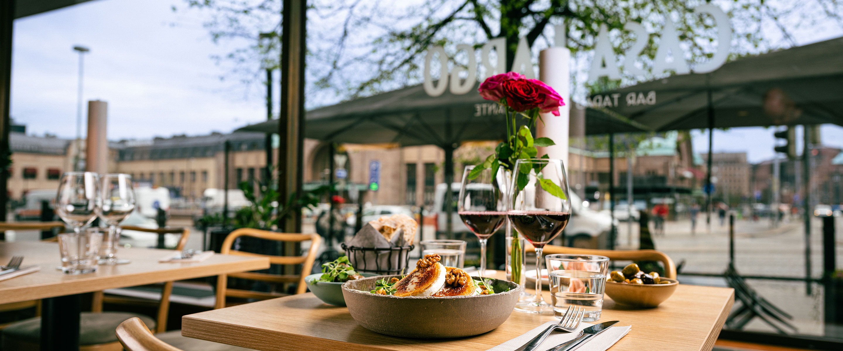 Restaurant table by glass windows featuring food, wine glasses and fresh roses in a vase.