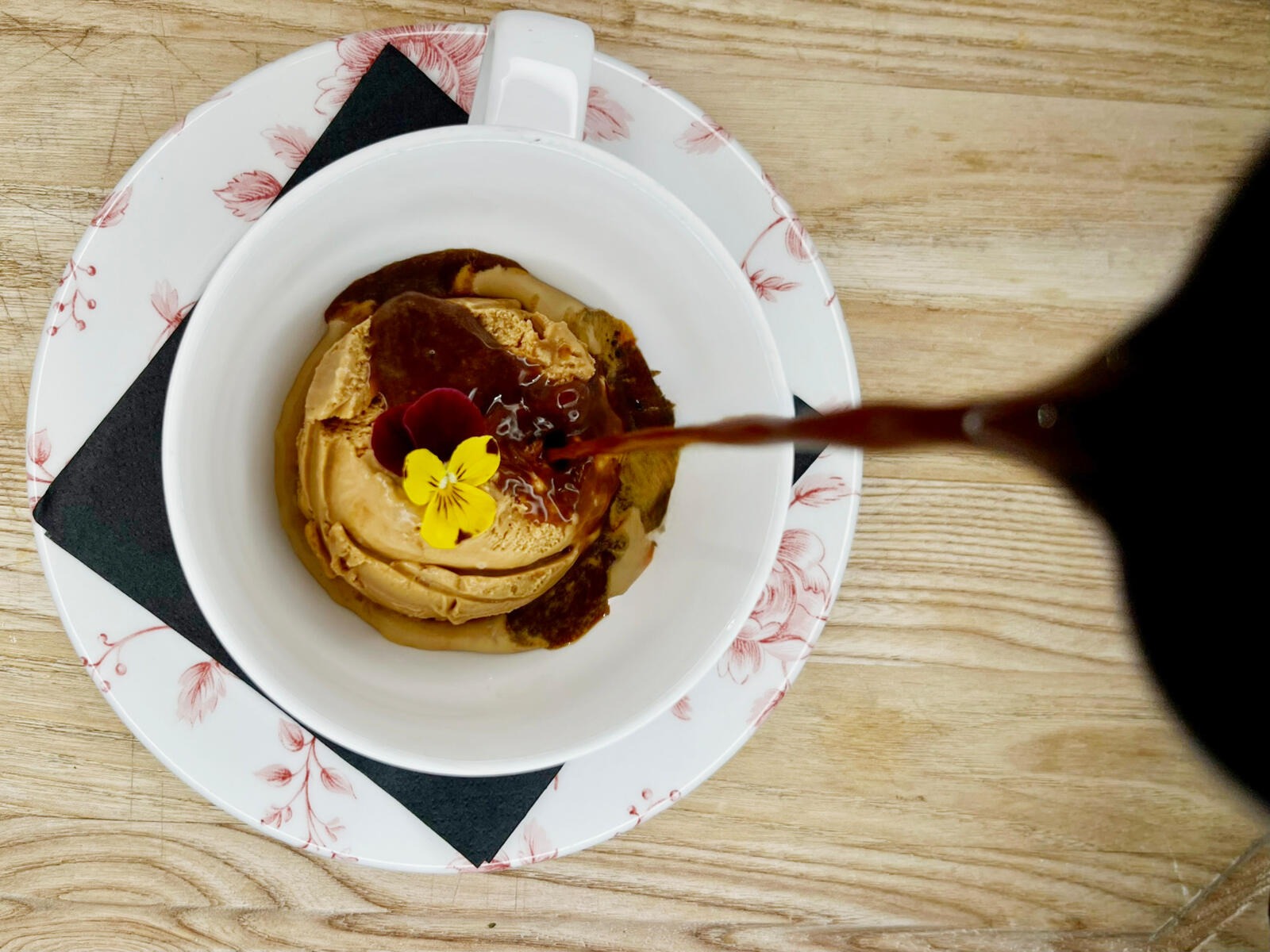 Dessert in a white bowl featuring ice cream or pudding with caramel sauce and garnished with a yellow edible flower.