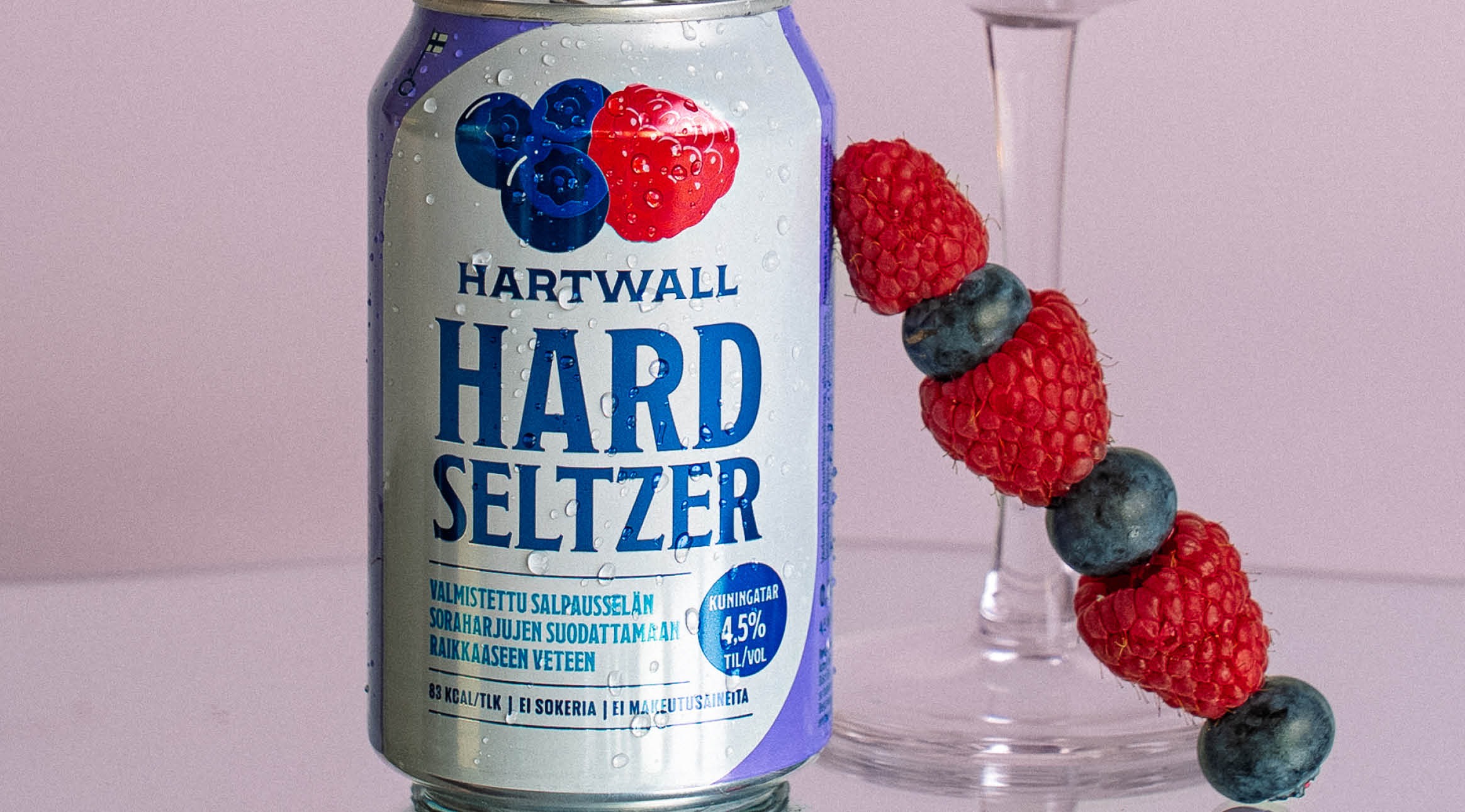 Hartwall Hard Seltzer -tölkki, jossa marjoja kuvituksena. Vieressä lasissa vartaassa vuorotellen vadelmia ja mustikoita.