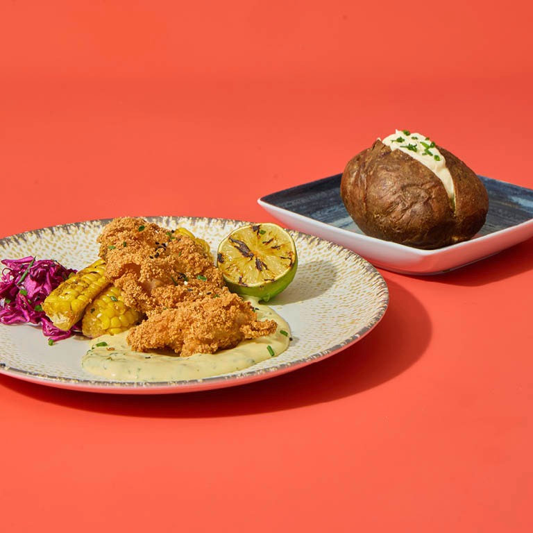 Ateria oranssilla taustalla: paneroitua broileria, maissia, limettilohko ja punakaalia lautasella, vieressä peruna.