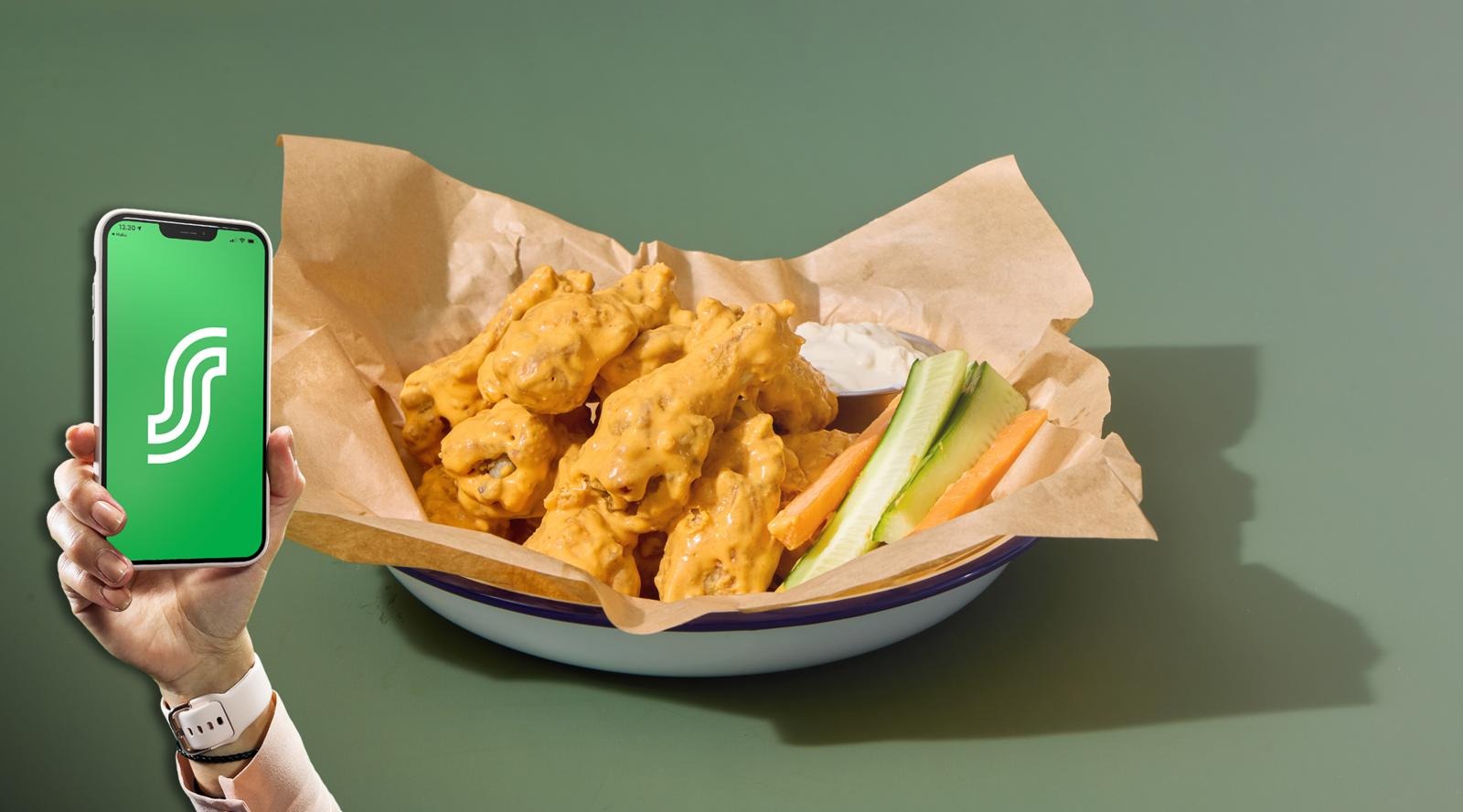 A hand holding a smartphone with green screen, beside a bowl containing fried food, carrot and cucumber sticks against a green background.
