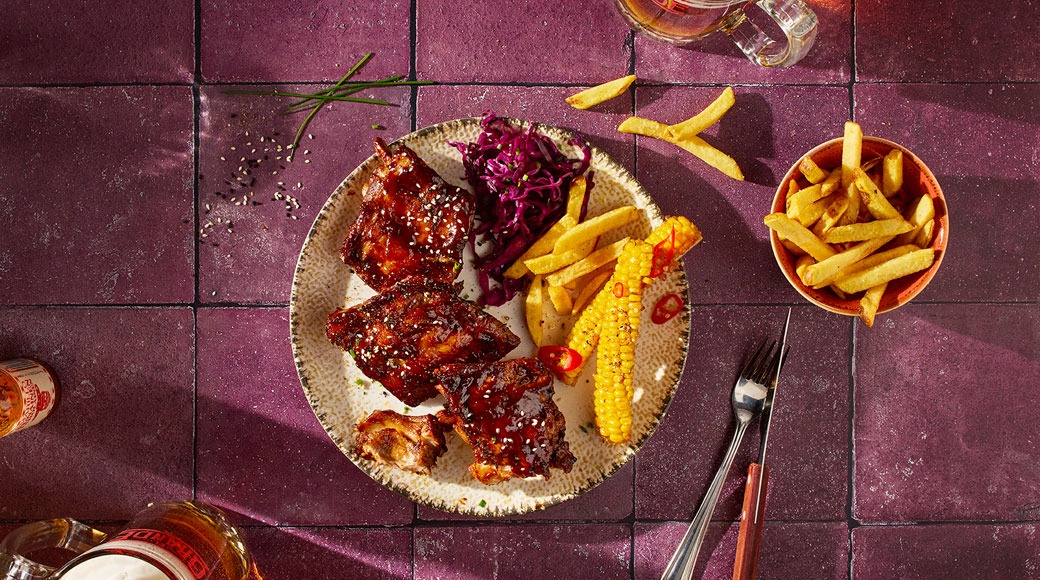 Glasuuritud grillitud ribid, friikartulid, mais ja punase kapsa salat taldrikul lillal plaatlaual. Kõrval on kahvel, nuga ja õlleklaasid.
