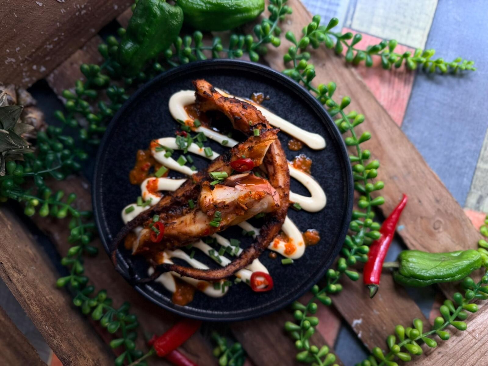 Black plate with grilled chicken on white sauce, garnished with green onions and chili slices, surrounded by fresh herbs and chili peppers on wooden table.