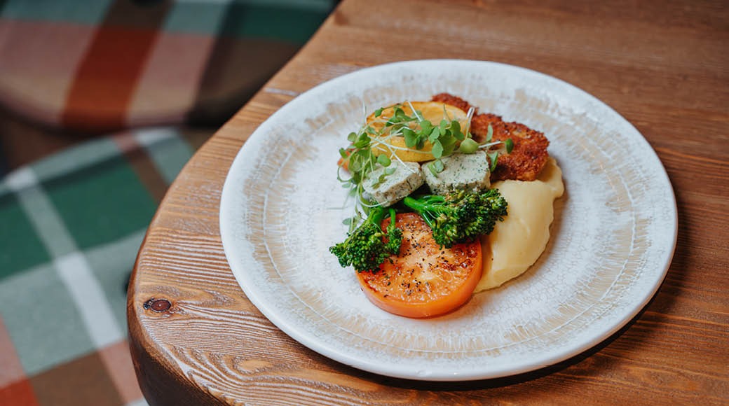 Lautasella tarjoiltu annos, jossa on perunamuusia, paistettua tomaattia, parsakaalia ja mikroversoja puisella pöydällä.