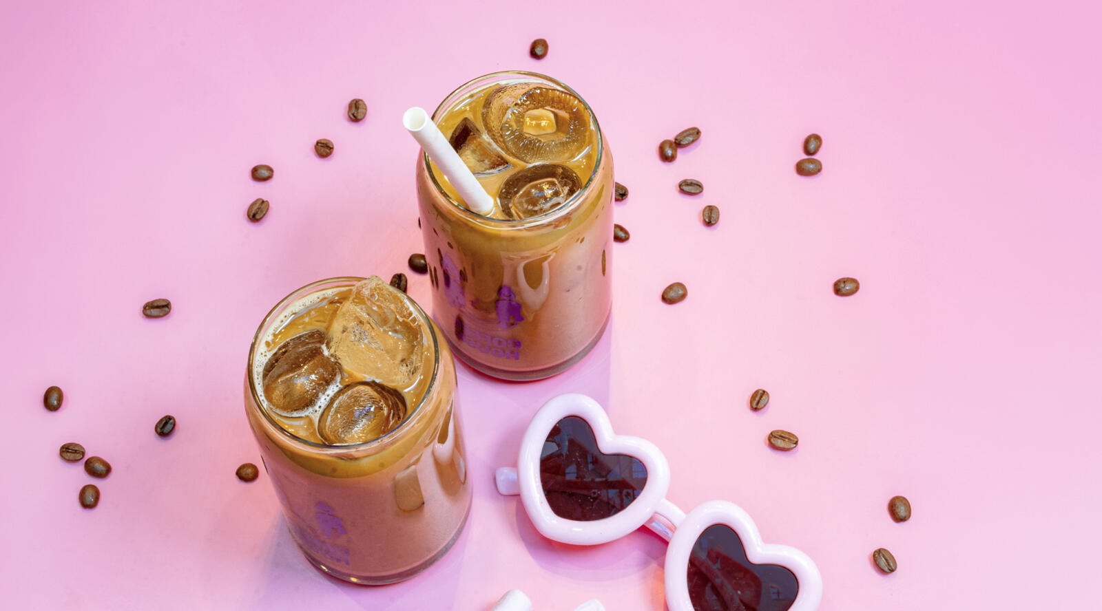 Två glas iskaffe med sugrör på rosa bakgrund, omgivna av kaffebönor och hjärtformade isbitar.