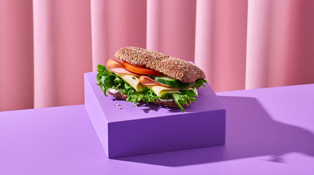 Täysjyväleipä, jossa on kinkkua, juustoa, tomaattia, kurkkua ja salaattia, violetilla alustalla vaaleanpunaista verhoa vasten.