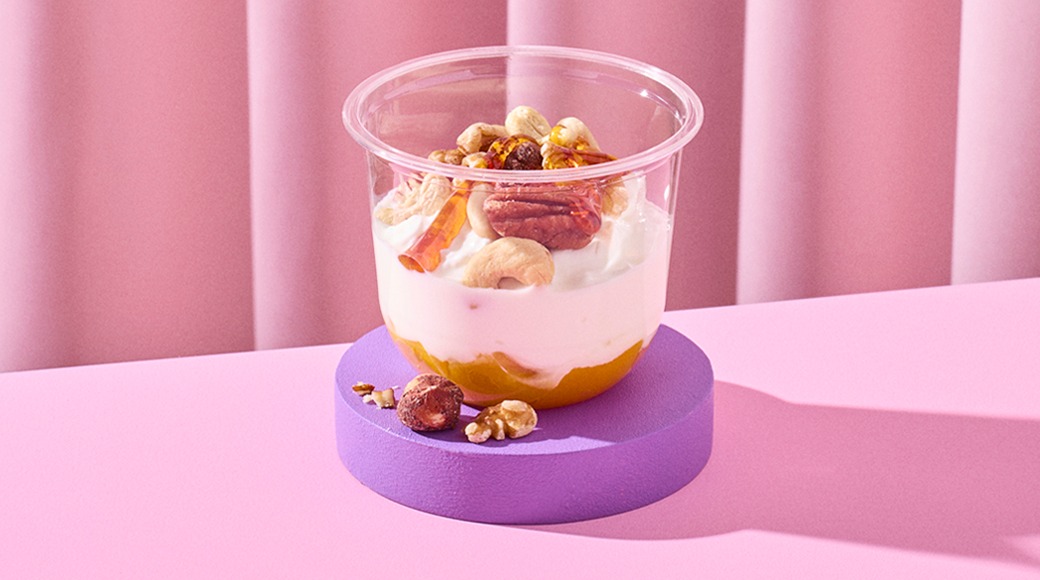 Kirkkaassa kupissa jogurttia, hedelmäsosetta ja pähkinöitä, kuten cashewpähkinöitä ja pekaanipähkinöitä. Kuppi on violetilla alustalla vaaleanpunaista taustaa vasten.