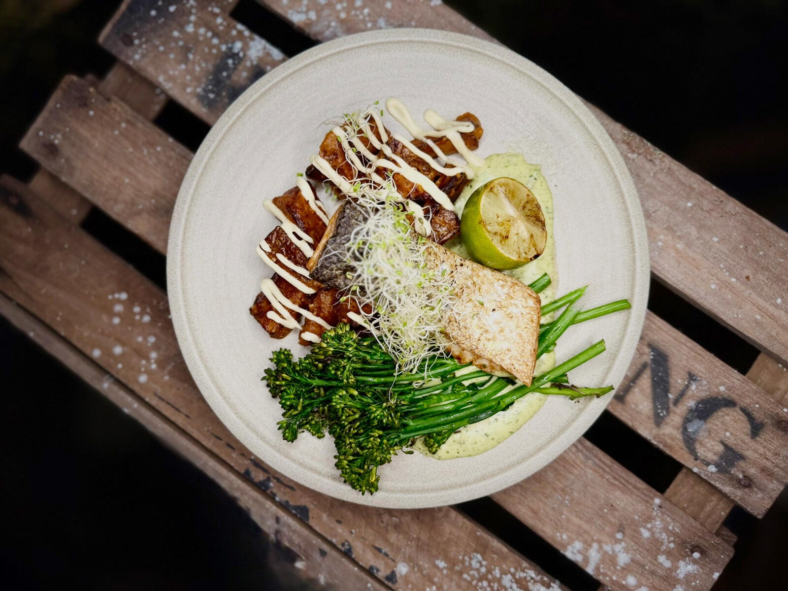 White plate on wooden surface featuring fish dish, roasted potatoes, broccolini, and a lime slice.