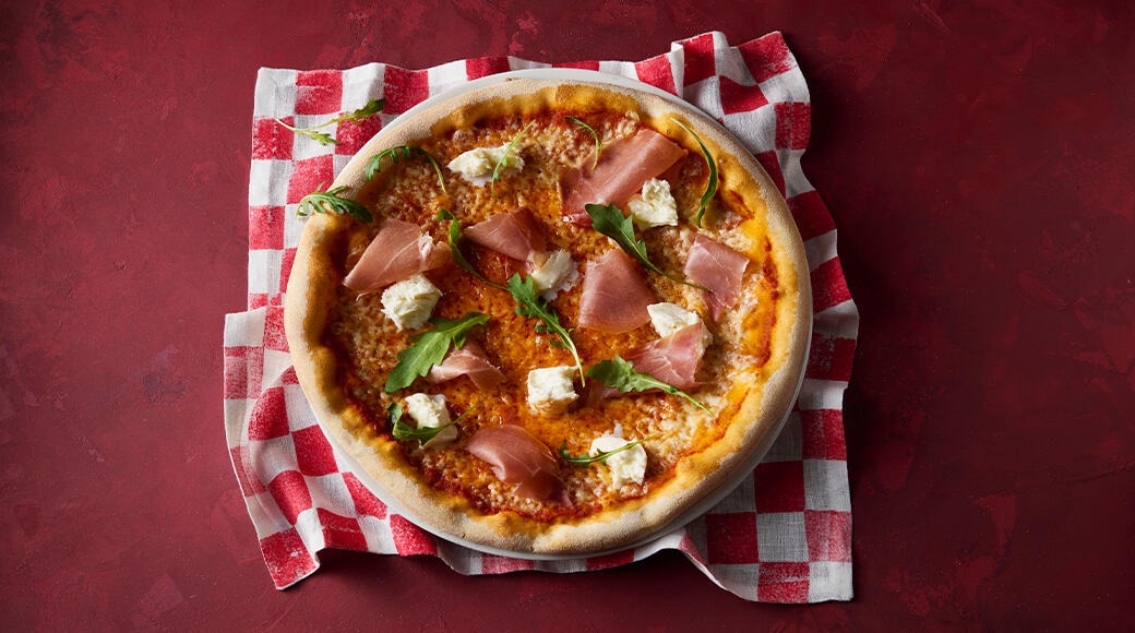 Pizza topped with prosciutto, fresh rocket and mozzarella. The pizza sits on a red and white checkered cloth on a red table.