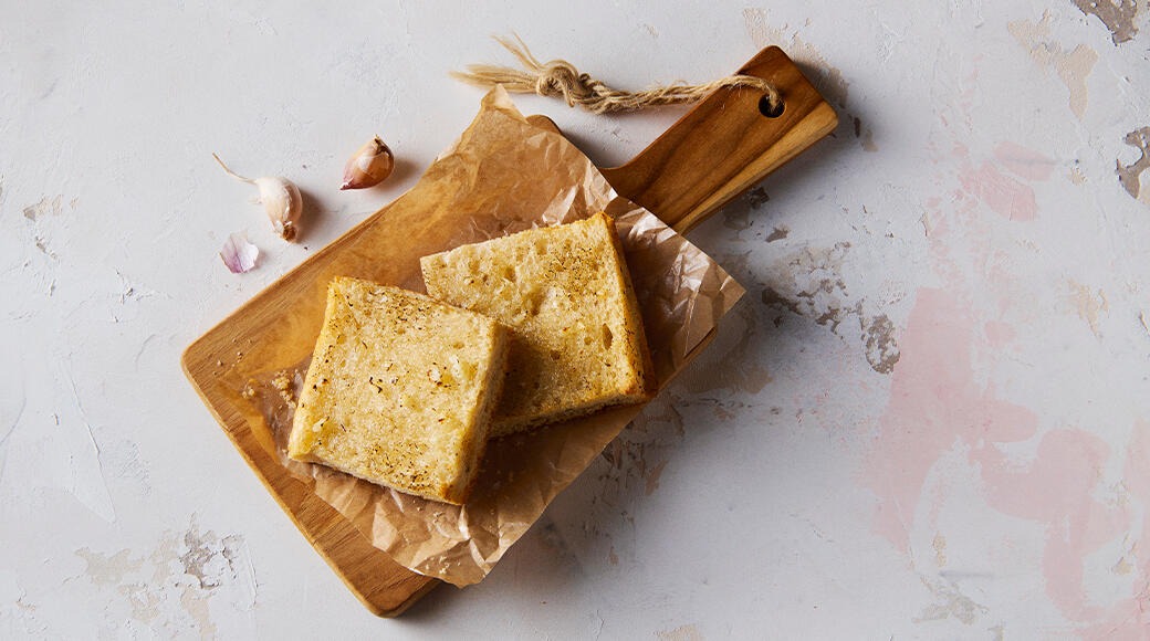 Two slices of garlic bread on baking paper on a wooden cutting board. Garlic cloves lie beside it on a light surface.