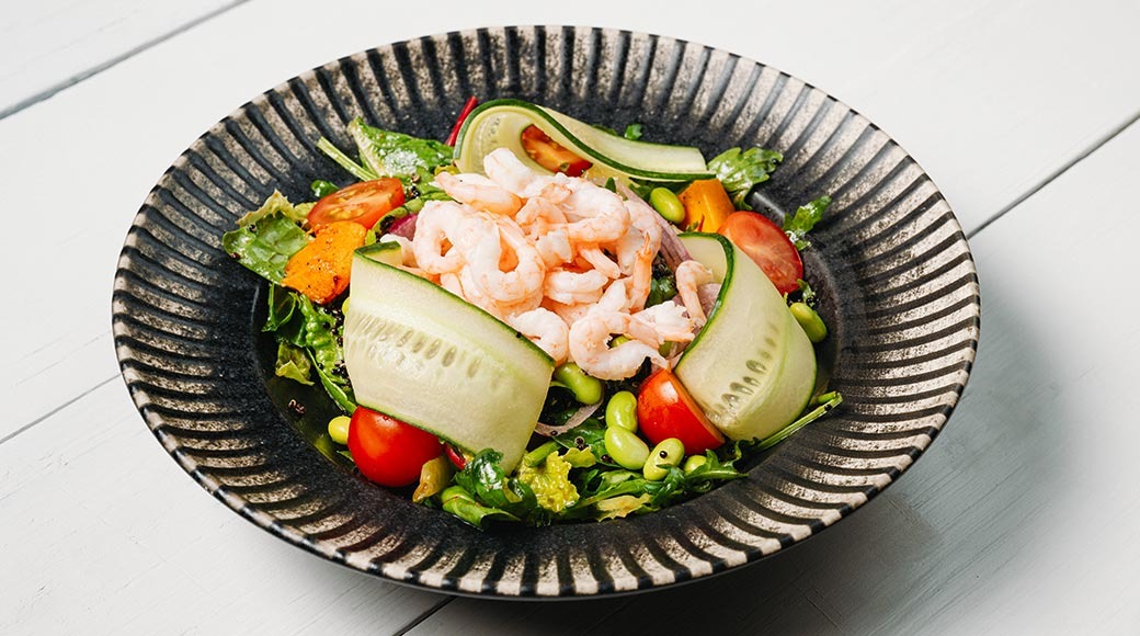 Krevetisalat tumedas ribilises kausis valgel laual. Peal kurgilindid, tomatid, edamame oad ja rohelised lehed.