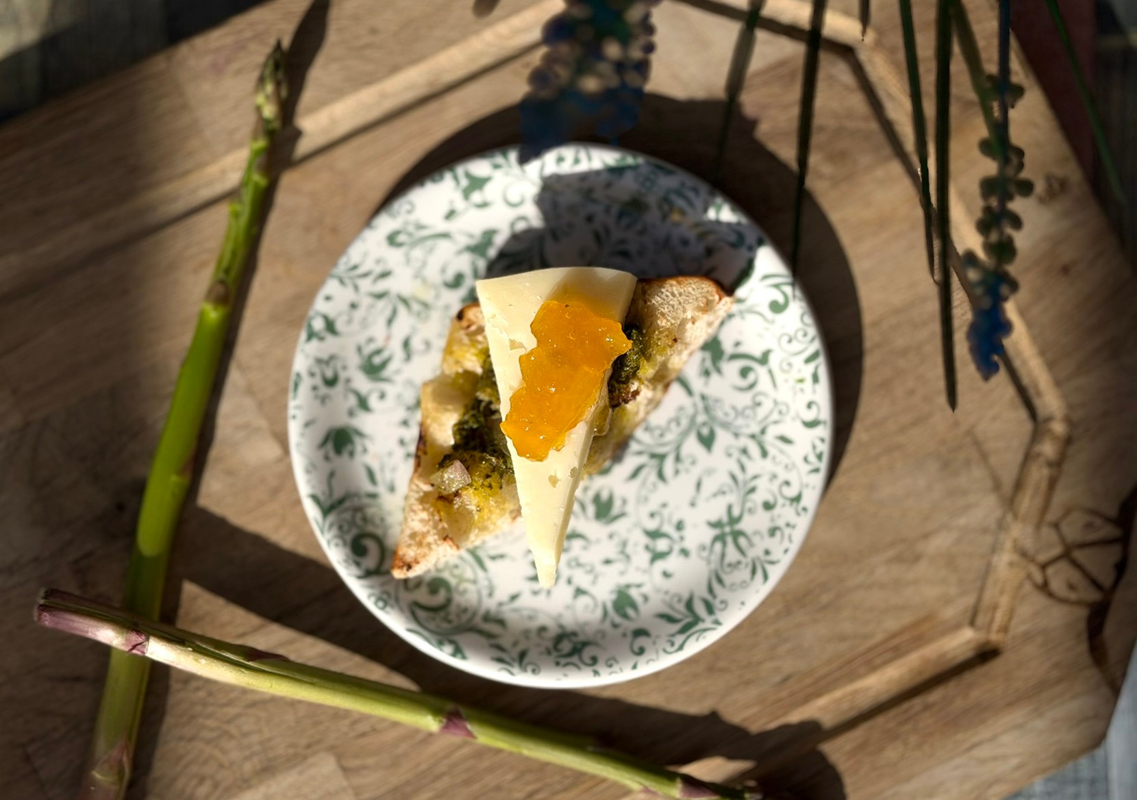 Cheese slice with jam on bread, served on a green patterned plate, on a wooden tray surrounded by decorative stems.