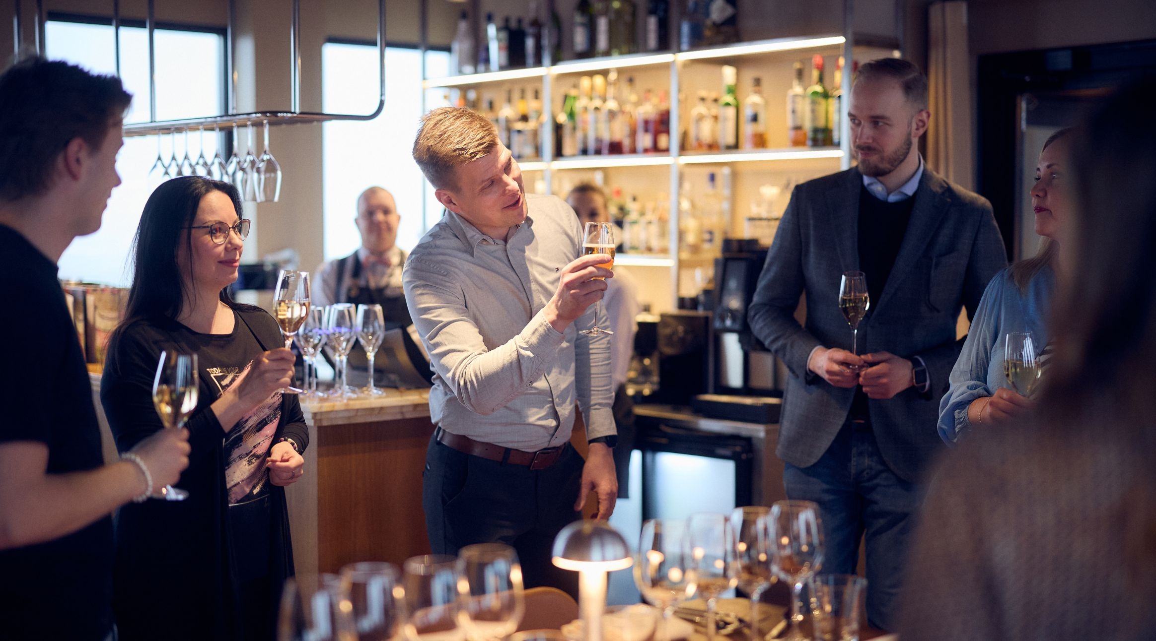 Ryhmä ihmisiä nauttimassa juomia tyylikkäässä baarissa, taustalla valaistut hyllyt täynnä pulloja.