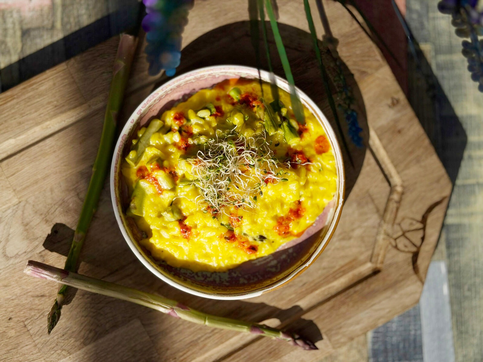 Yellow risotto-like dish in a bowl on wooden base, garnished with sprouts and asparagus stems around it.