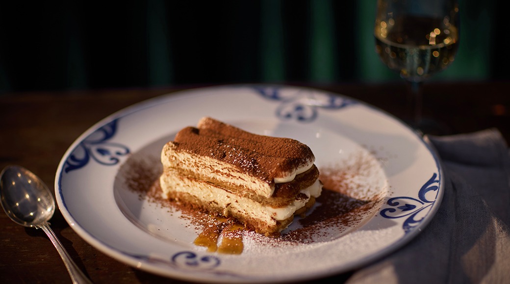Tiramisu-dessert på en vit tallrik med blå dekorationer. Ett vinglas syns i bakgrunden.