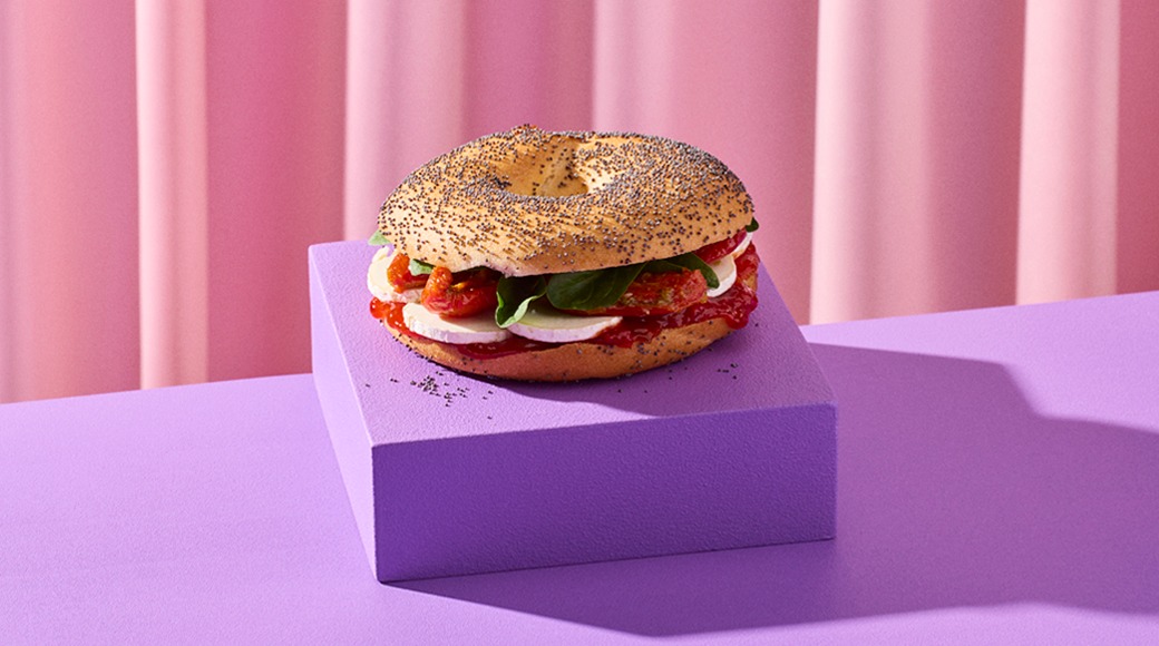 Unikonsiemenillä ripoteltu bagel, jonka välissä on mozzarellaa, aurinkokuivattuja tomaatteja ja pinaatinlehtiä. Bagel on violetin korokkeen päällä vaaleanpunaista verhoa vasten.