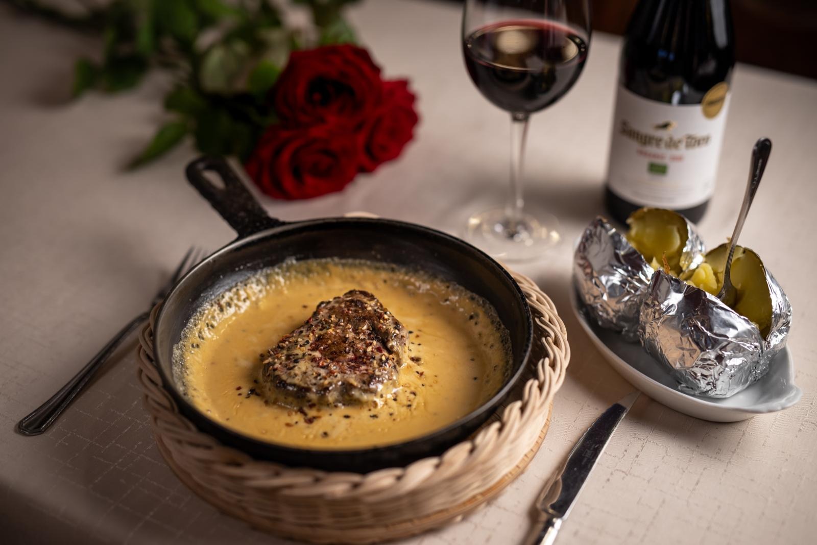 Pihviannos kastikkeessa pannulla, vieressä folioperunat, punaviinilasi, viinipullo ja punaisia ruusuja romanttisessa kattauksessa.