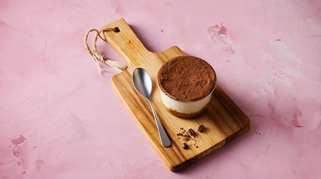 Tiramisu dusted with cocoa in a glass dish on a wooden serving board. A spoon and coffee beans lie beside it on a pink background.
