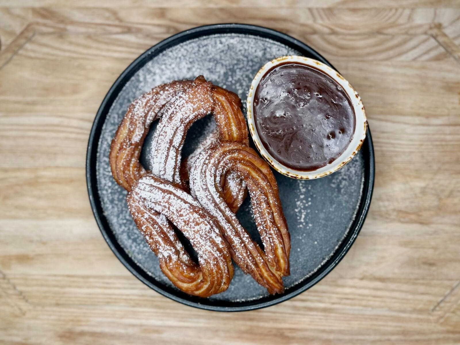 Sugar-dusted churros with chocolate dipping sauce on a gray plate atop wooden table.