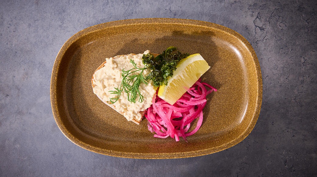 Ruskealla lautasella kalatahnaa, vihreää kastiketta, sitruunalohko ja pinkki sipulisalaatti harmaalla pohjalla.