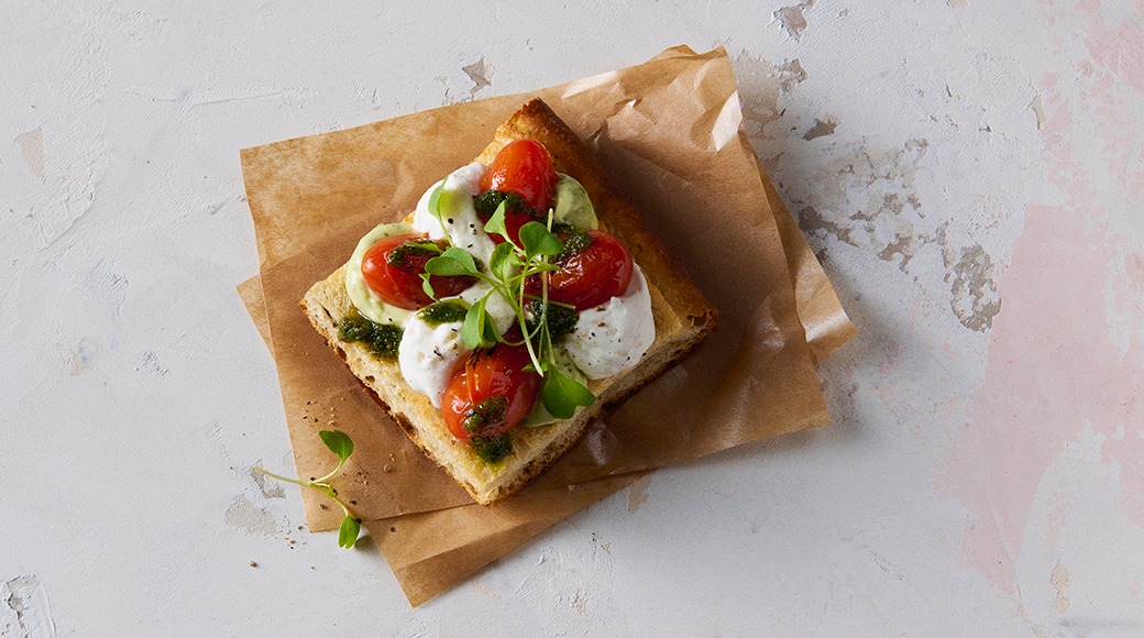 Square slice of bread topped with tomato halves, soft cheese, pesto and microgreens on baking paper.