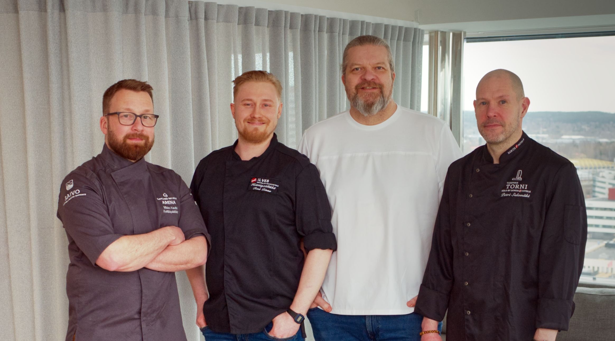 Four people standing in a row in front of a window with light curtains, three wearing chef uniforms and one in a white shirt and jeans.