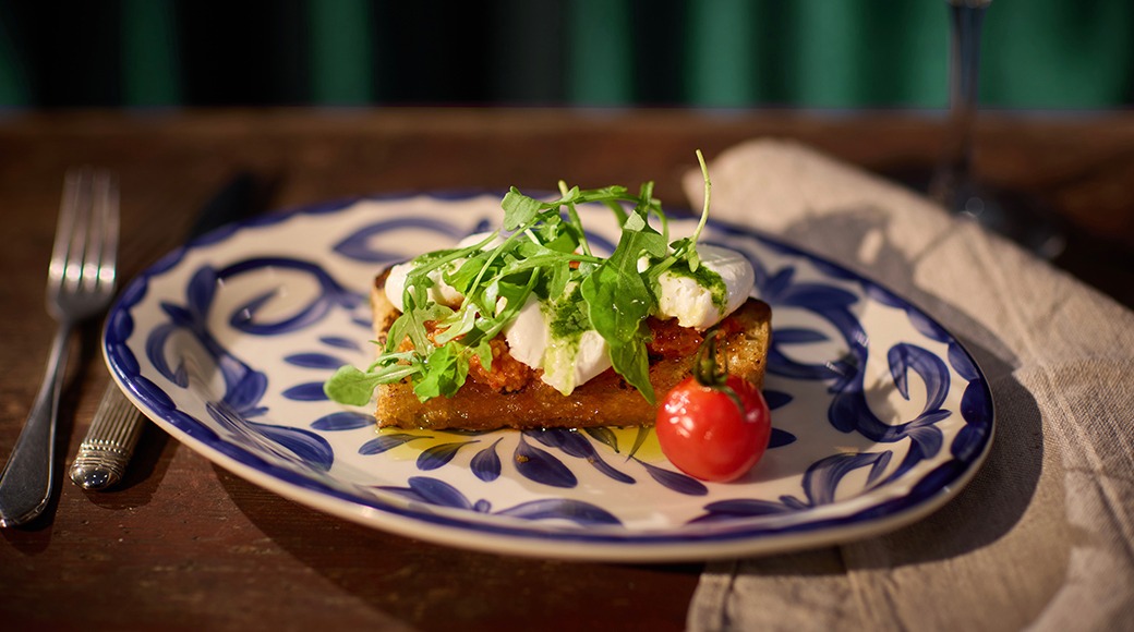 Bruschetta med getost och rucola på tallrik. En körsbärstomat bredvid. Dekorativ blå-vit tallrik på träbord.