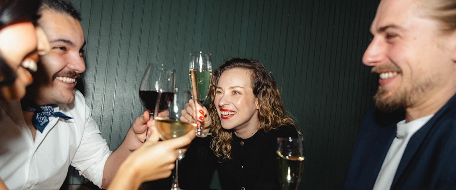 Four people toasting with wine glasses in front of a dark green wall, smiling.