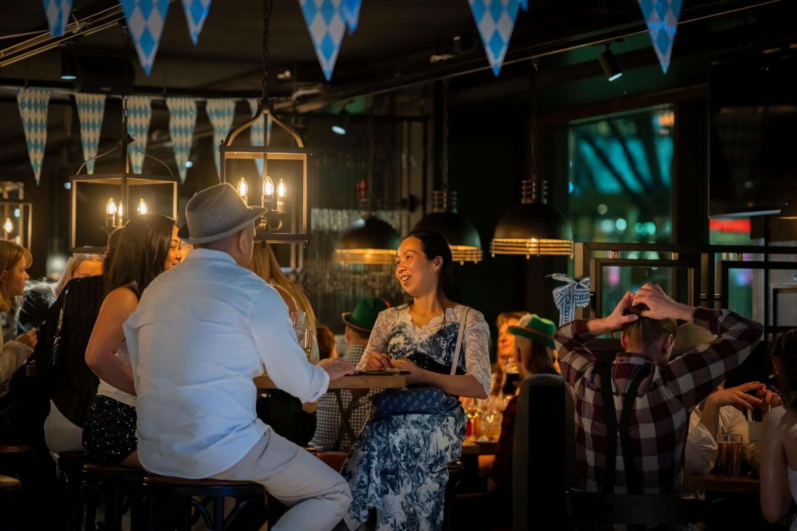 Oktoberfest-tunnelmaa ravintolassa, jossa siniset ja valkoiset viirit koristavat kattoa. Asiakkaat keskustelevat pöytien ääressä, osa on pukeutunut teeman mukaisesti.