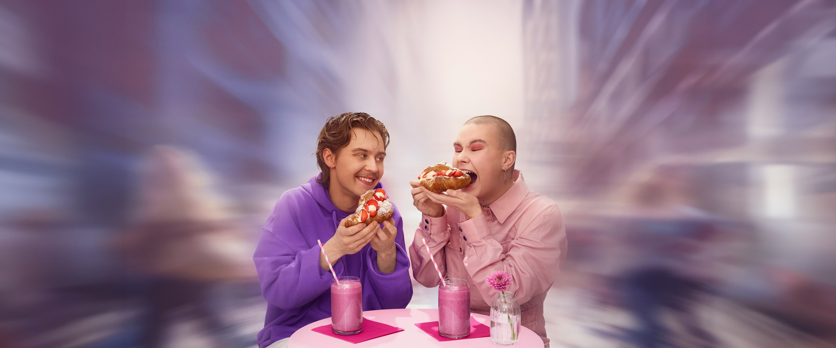 Kaksi henkilöä maistavat täytettyä croissanttia. Pöydällä kaksi smoothiekuppia.