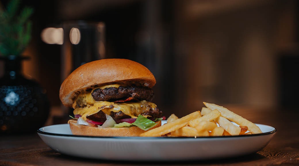 Mehevä hampurilainen, jossa kaksi pihviä ja sulatejuustoa, tarjoiltuna ranskanperunoiden kanssa valkoisella lautasella.