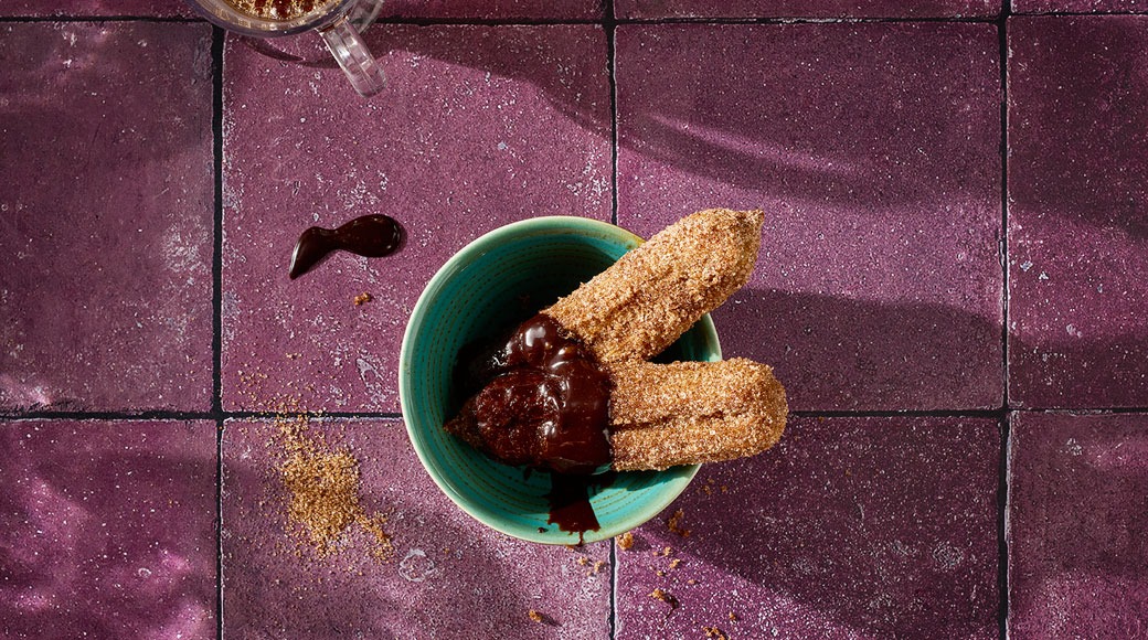 Kaks suhkrustatud churrost ja šokolaadikaste türkiissinises kausis lillal plaaditud pinnal. Kõrval on kaneelisuhkrut ja šokolaaditriip.