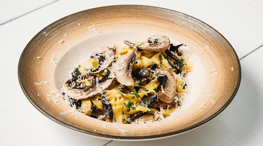 Tagliatelle seentega, riivjuustu ja ürtidega pruunide servadega sügaval taldrikul valgel laual.