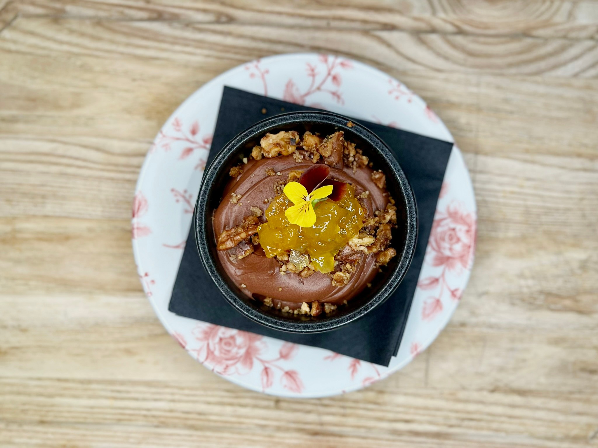 Chocolate dessert in a black bowl garnished with yellow edible flower, fruit preserves and nut crumble, served on a decorative plate on wooden table.