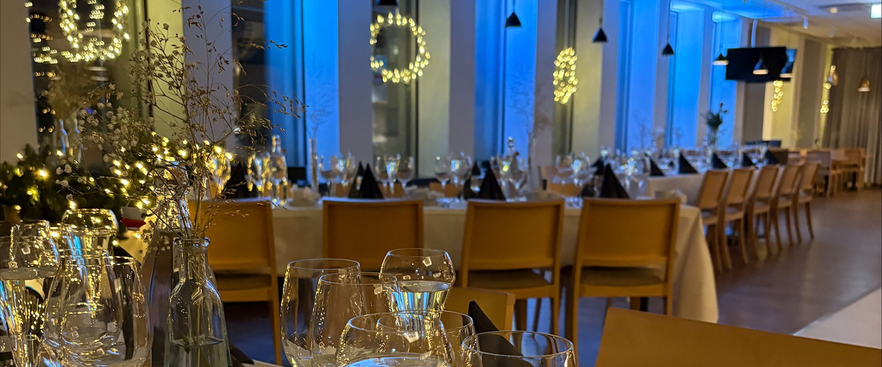 Ravintolan juhlatila, jossa pitkiä pöytiä, sinertävä valaistus ja koristevaloja. Etualalla laseja ja maljakoita.