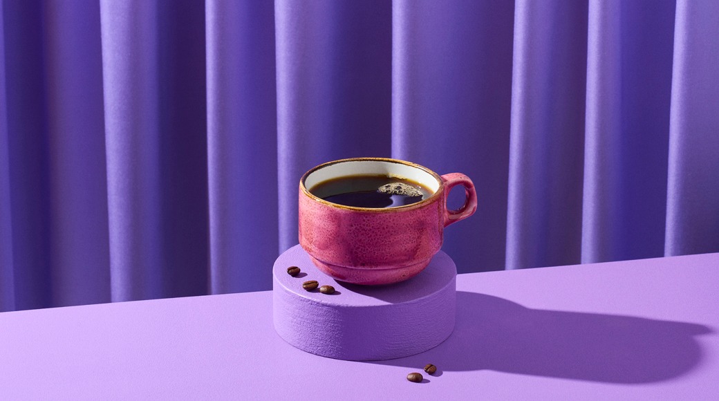 Pinkki keraaminen kuppi mustaa kahvia violetin korokkeen päällä. Ympärillä on kahvipapuja, taustalla violetti verho.