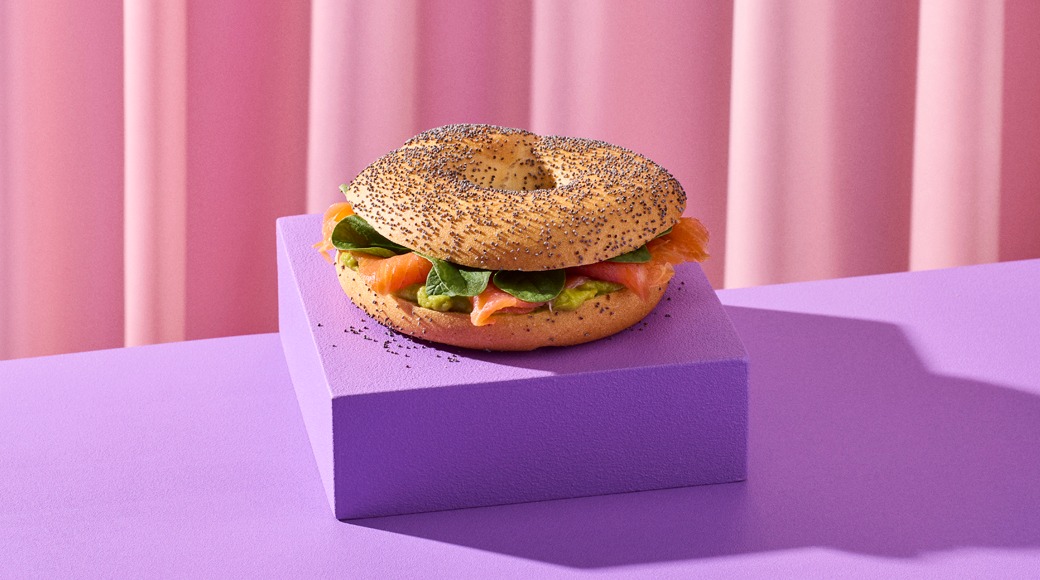 Unikonsiemenillä ripoteltu bagel, jonka välissä on savulohta, pinaatinlehtiä ja avokadoa. Bagel on violetin korokkeen päällä vaaleanpunaista verhoa vasten.