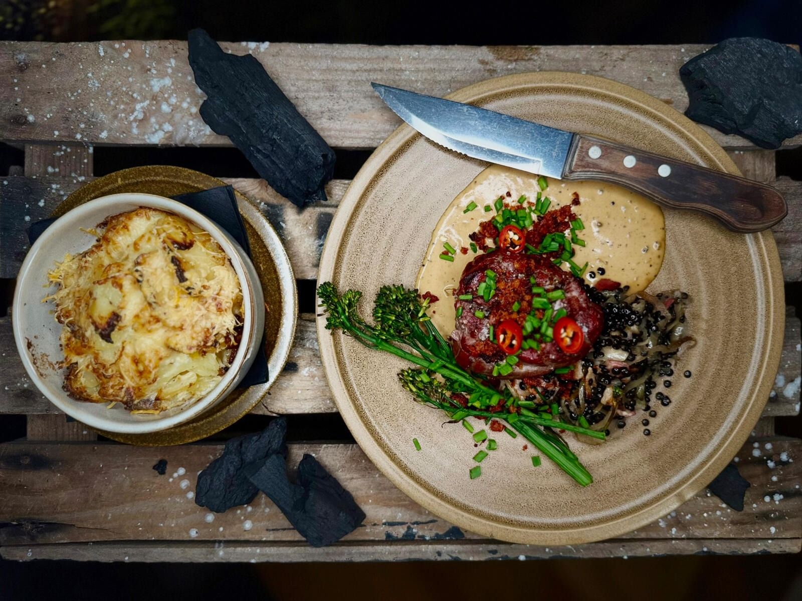 Two dishes on wooden surface: a small bowl with gratinated food and a plate with steak tartare garnished with green herbs and sauce, knife alongside.
