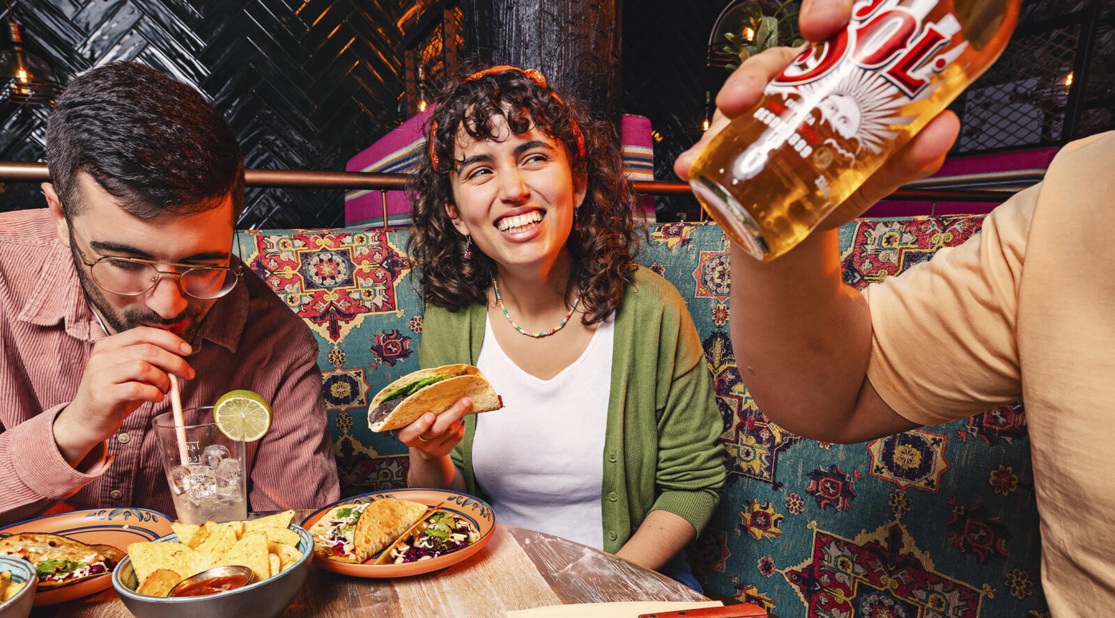 Kolm inimest restoranis naudivad mehhiko toitu, keskel naine naeratab hoides tacot. Laual on nachod ja joogid.